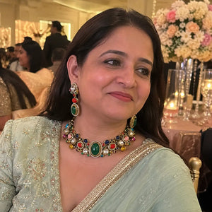 A woman wearing a light sage green embroidered traditional outfit and a prominent multicolor gemstone necklace with matching drop earrings, photographed in a decorated indoor event setting.